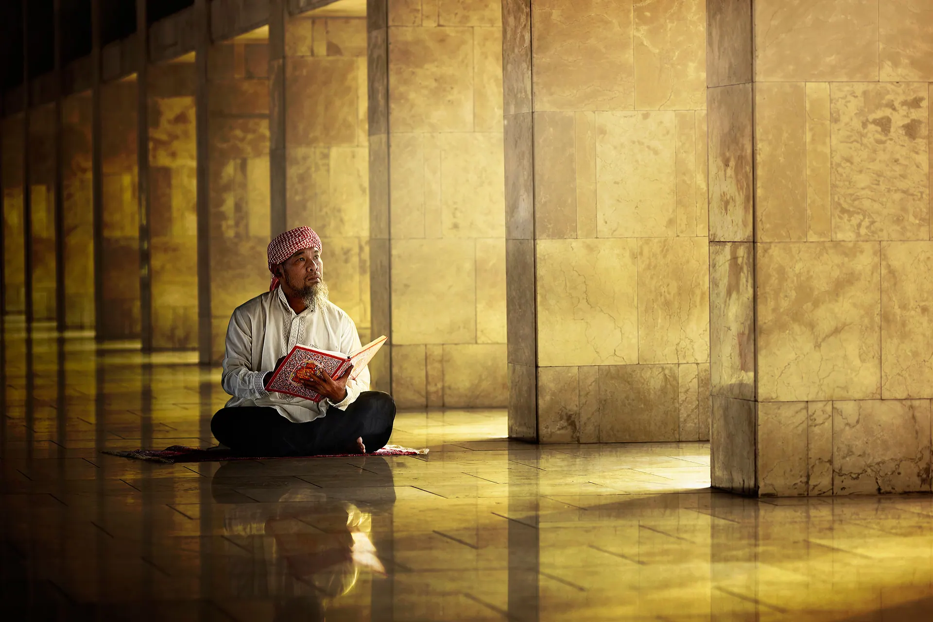 A man reading the Qur’an seated on a prayer mat in a golden-lit mosque colonnade